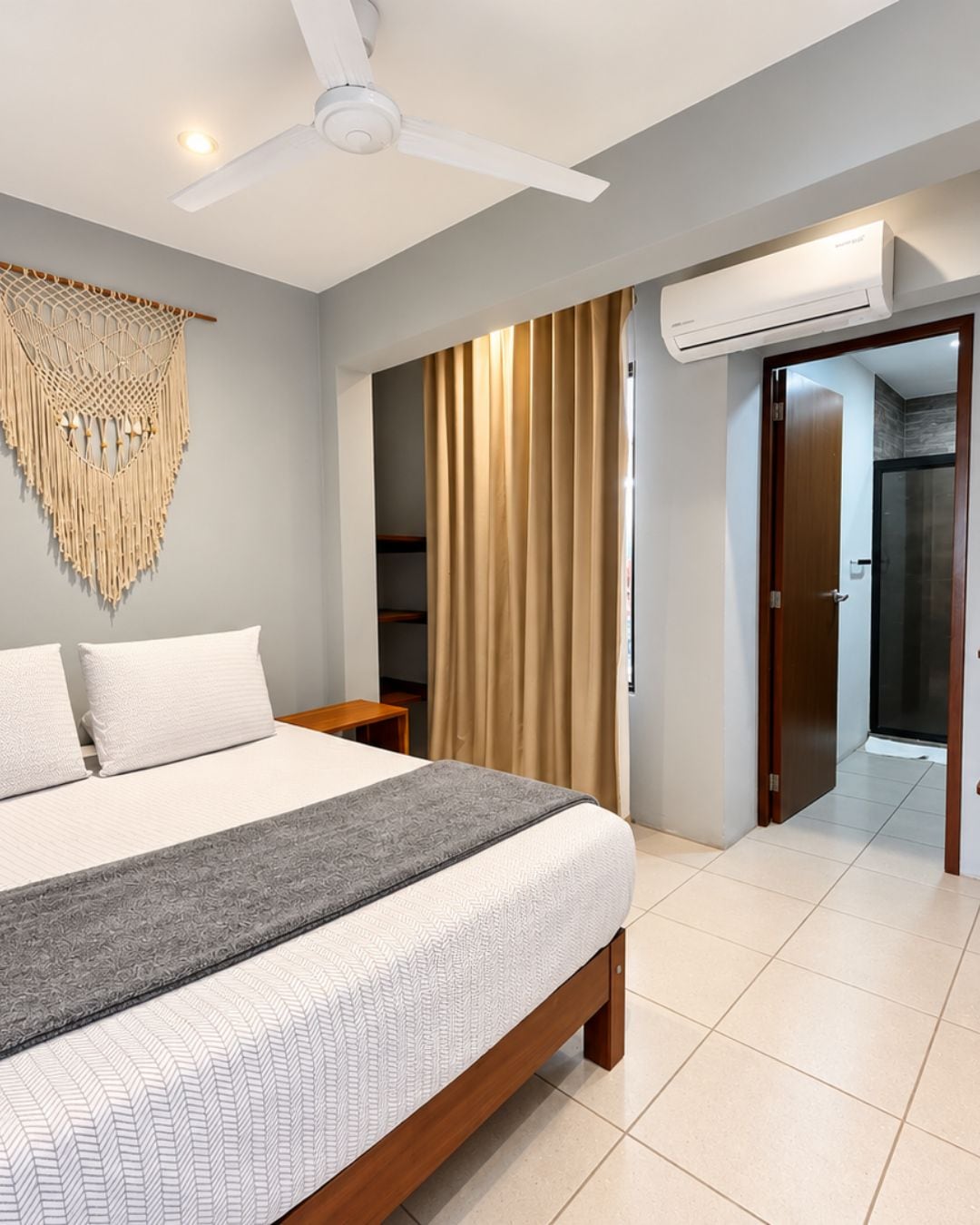 Une chambre avec un lit, un ventilateur de plafond, une décoration murale en macramé, une fenêtre avec des rideaux et un appareil de climatisation.