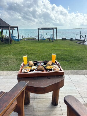Une table en bois, dressée avec des aliments et des boissons pour le petit-déjeuner, donnant sur une zone herbeuse et un plan d’eau.
