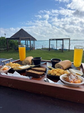 Un plateau de petit-déjeuner avec des œufs, des toasts, des fruits et du jus, disposé devant une vue sur la mer et un belvédère.