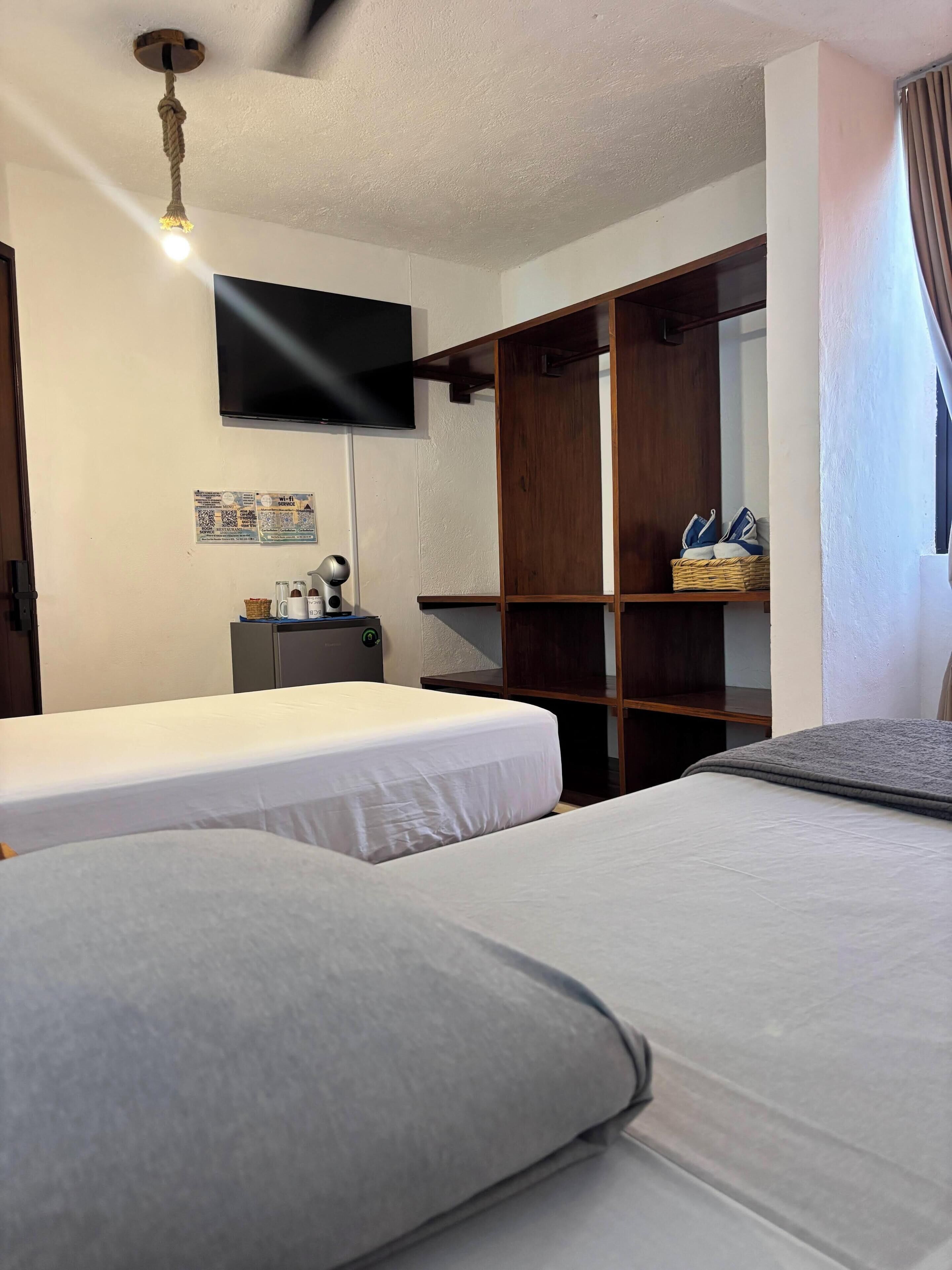 Habitación de hotel con dos camas, un televisor montado en la pared, un armario de madera y una mesita de noche con lámpara.