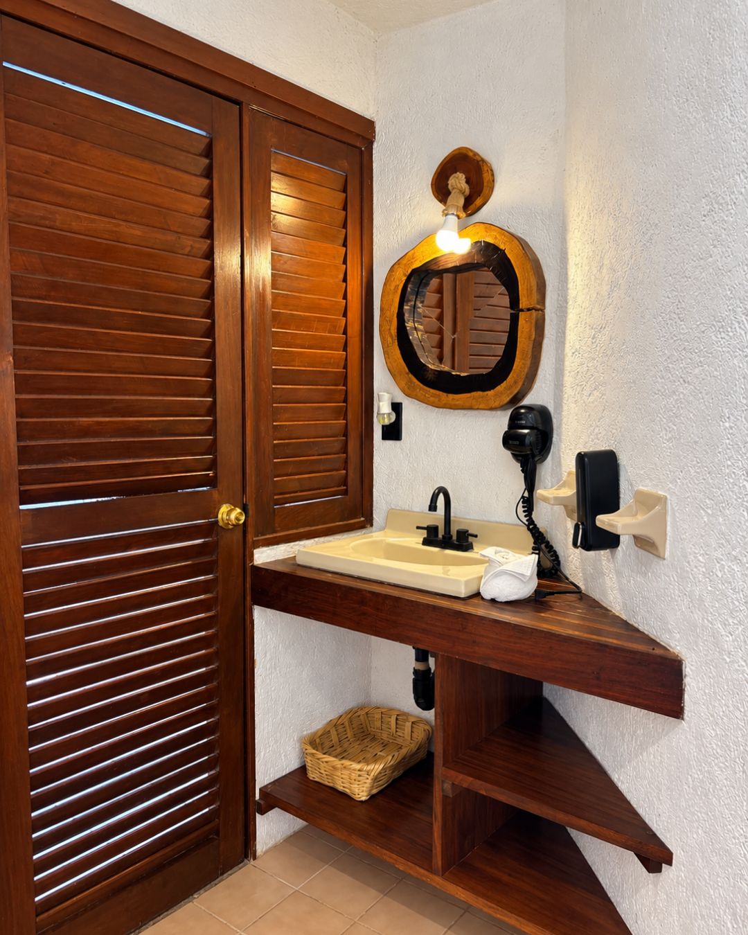 Un baño con puerta de madera, un lavabo con espejo, una toalla, una cesta y un teléfono en la pared.