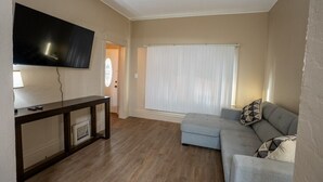 Un salón con un televisor de pantalla plana montado en la pared, una consola de madera oscura, un sofá gris con cojines estampados y un amplio ventanal con persianas blancas.