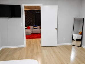 Une pièce avec un plancher en bois, une porte blanche, un téléviseur à écran plat fixé au mur et un lit recouvert d’une couverture blanche.