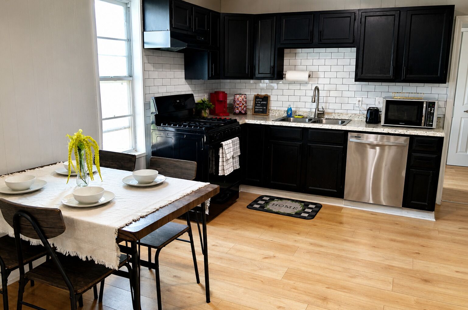 Una cocina moderna con armarios negros, un lavavajillas de acero inoxidable y un comedor con mesa dispuesta para cuatro.