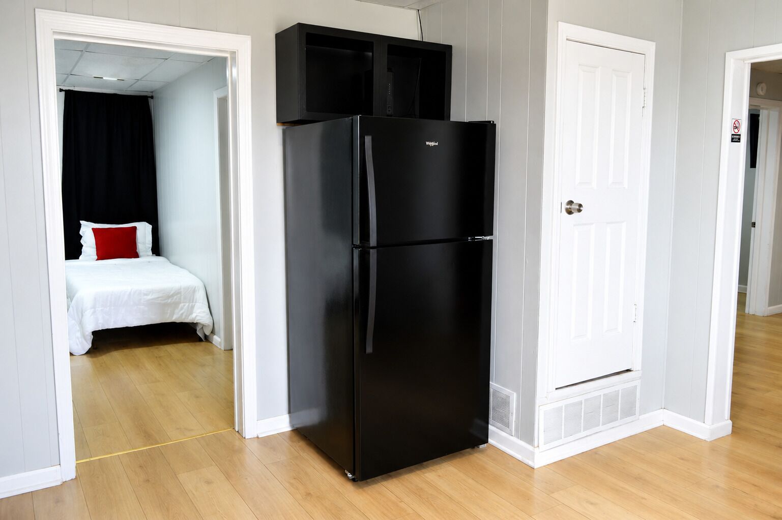 A compact room with a black refrigerator, a bed with white bedding and a red pillow, and a white door with a 'No Entry' sign.