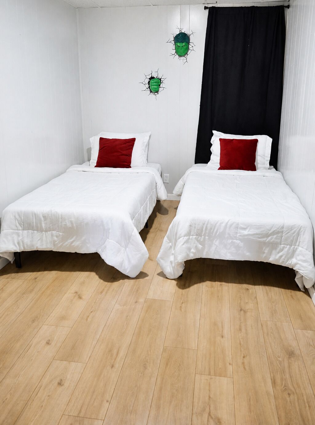 Dos camas individuales con ropa blanca y almohadas rojas, suelo de madera y cortina negra.