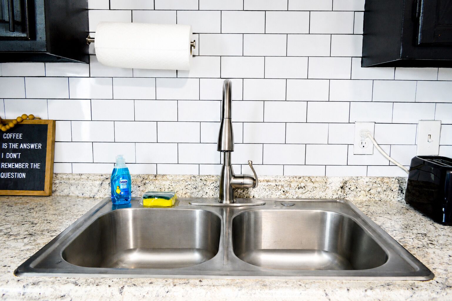 A kitchen with a double stainless steel sink, a roll of paper towels, a bottle of dish soap, and a sponge on the counter.