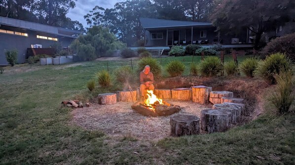 A backyard with a fire pit surrounded by tree stumps, a grassy area, and a house in the background.