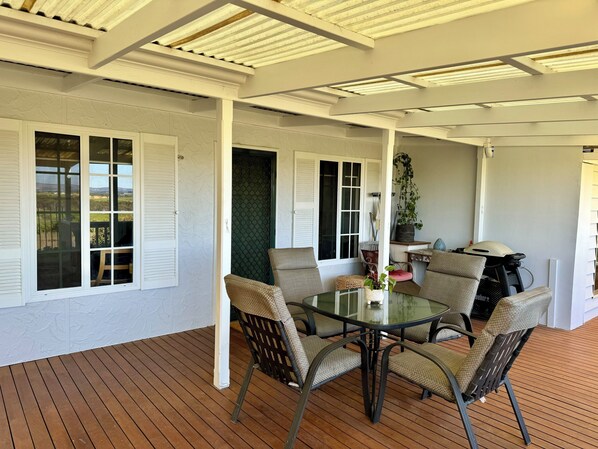 A covered outdoor dining area with a glass table and wicker chairs, surrounded by white walls and wooden flooring.