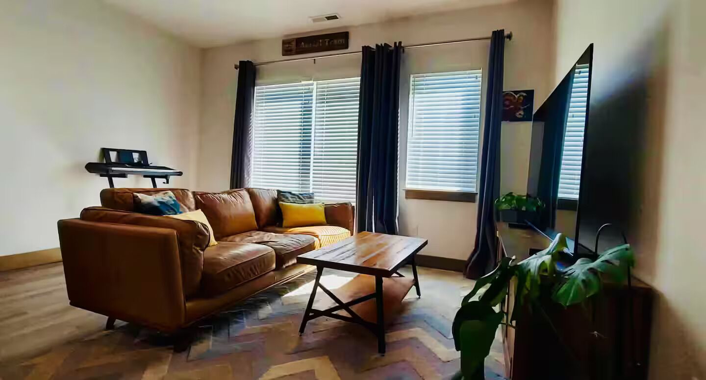 A living room with a brown leather sofa, a wooden coffee table, and a television. There are two windows with curtains, a sign above the sofa, and a plant in the corner.