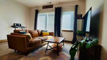 A living room with a brown leather sofa, a wooden coffee table, and a television. There are two windows with curtains, a sign above the sofa, and a plant in the corner.