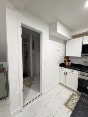 A kitchen with white cabinets, a black countertop, and a stove. There is a door leading to a bathroom with a shower. - Cozy Tampa Retreat Near Downtown, Riverwalk & Raymond James Stadium (Tampa)