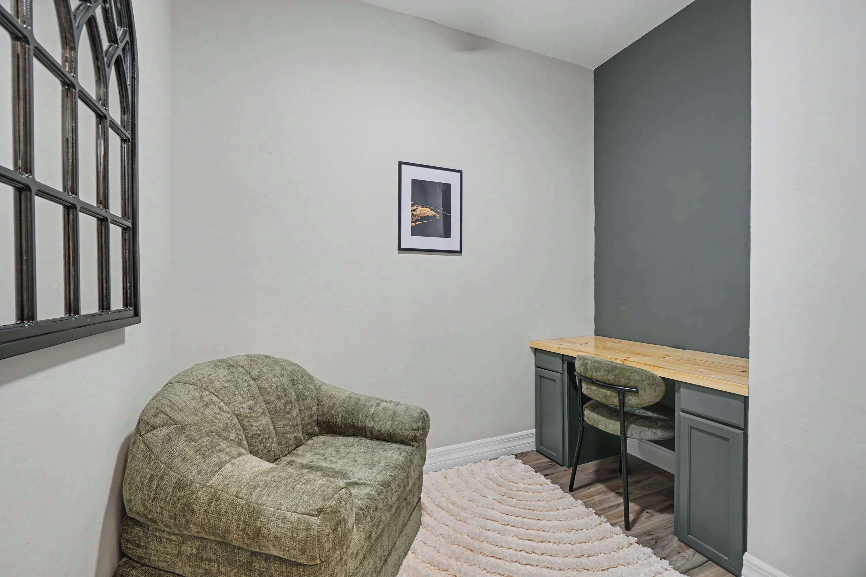 A corner room with a grey wall, a framed picture, a green armchair, a wooden desk, and a white shag rug.