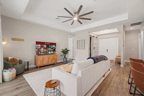 A modern living room with a sofa, a TV, a ceiling fan, and a wooden cabinet. - Cape Daydream | Heated Pool, Spa & Game Room (Cape Coral)