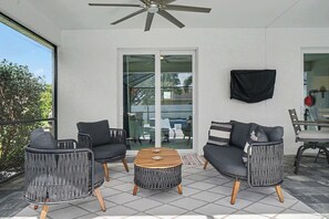 Ein überdachter Patio-Bereich mit Deckenventilator, Glastür zum Außenbereich und einer Sitzecke mit zwei Sesseln und einer Couch.