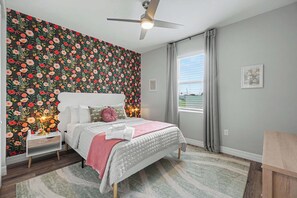 Un dormitorio con papel tapiz floral, una cama con cabecera blanca, una mesita de noche con lámpara y una ventana con cortinas.