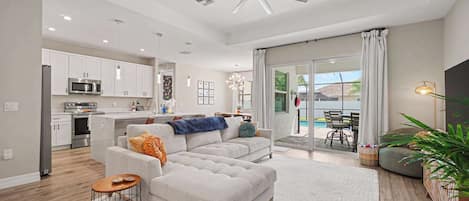 A modern living room with a grey sofa, a coffee table, and a dining area with a pool view.