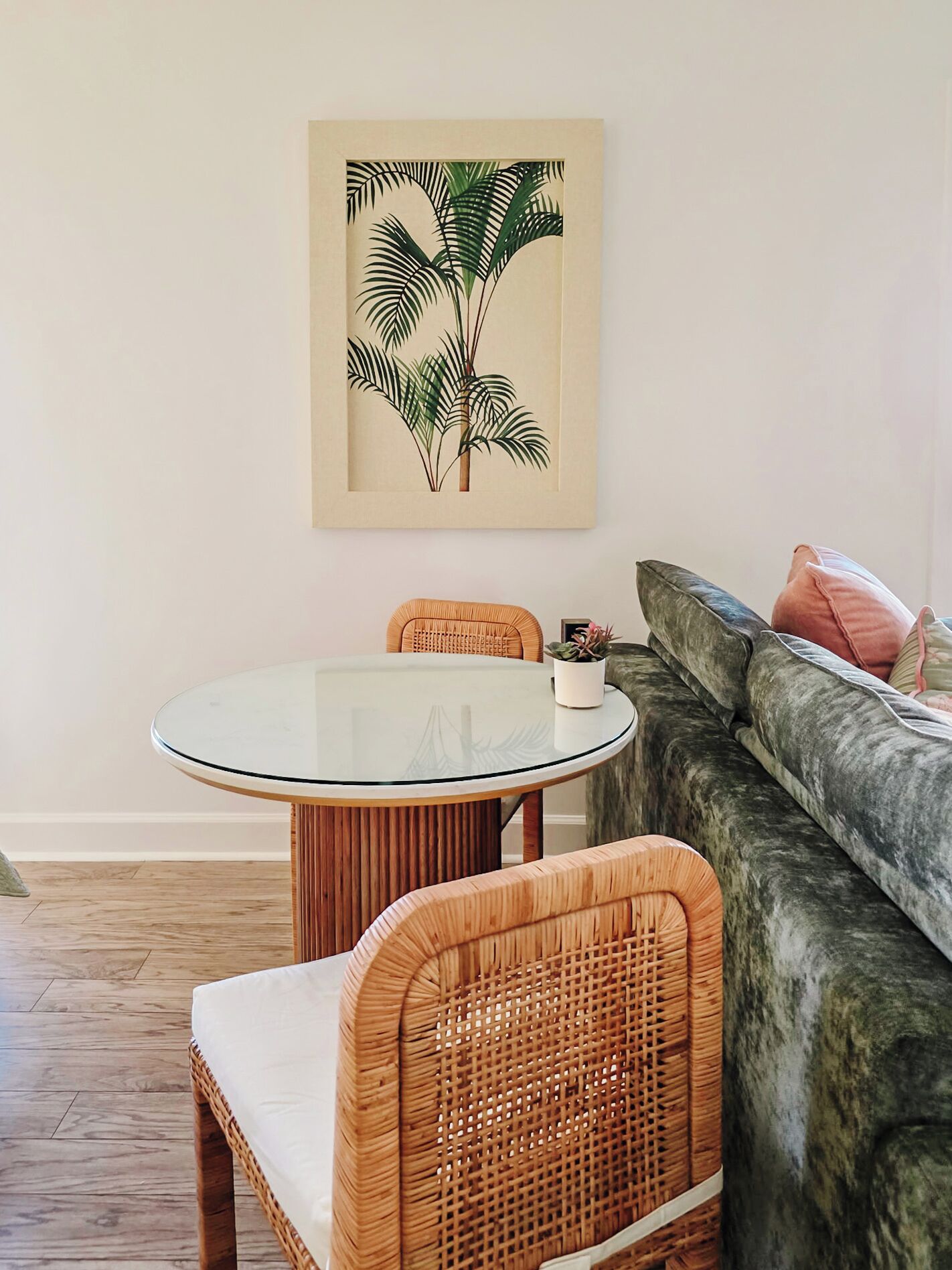 Een eetruimte met een glazen tafel, rieten stoelen, een gestoffeerde bank en een plantenillustratie aan de muur.