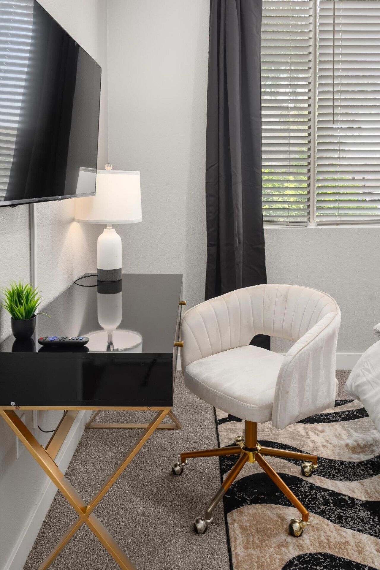Une chambre moderne avec un bureau noir, une chaise blanche, un téléviseur à écran plat, une lampe et une fenêtre avec des stores.