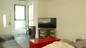 Uma sala de estar com uma televisão sobre um suporte de madeira, uma mesa de centro vermelha e um sofá.