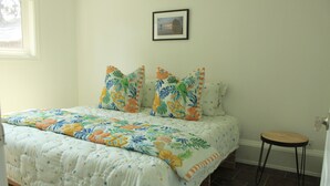 Quarto com cama, travesseiros, um banquinho, um quadro na parede e uma janela.