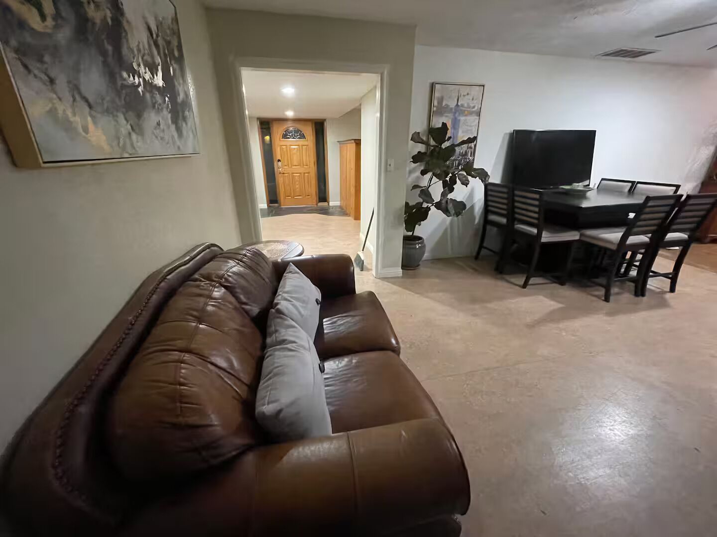 A living room with a brown leather sofa, a dining table with chairs, a potted plant, and a painting on the wall.
