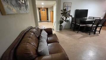 A living room with a brown leather sofa, a dining table with chairs, a potted plant, and a painting on the wall.