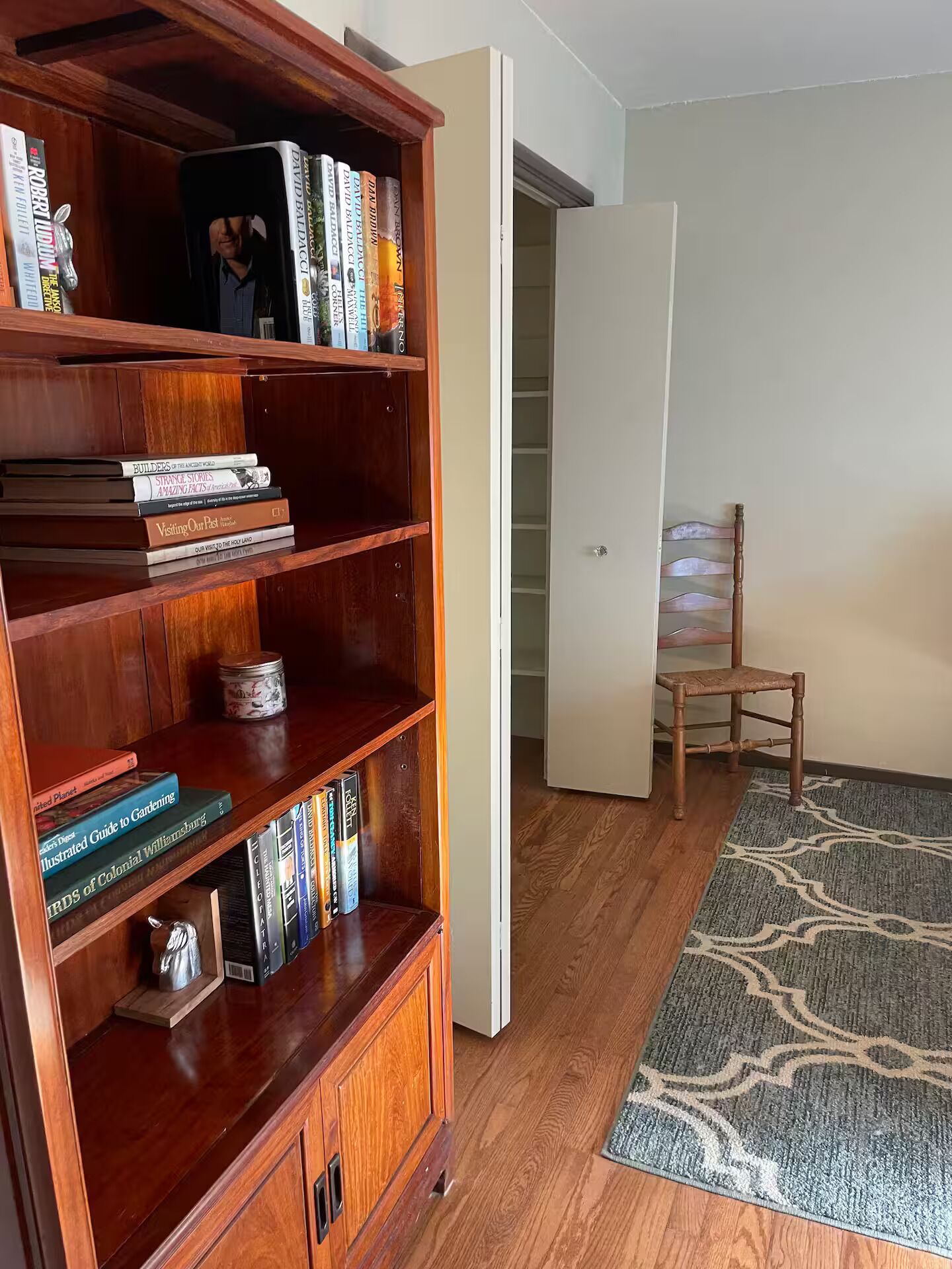 Ein Zimmer mit einem hölzernen Bücherregal, voll mit Büchern, einer weißen Tür, einem Holzstuhl und einem gemusterten Teppich auf dem Boden.