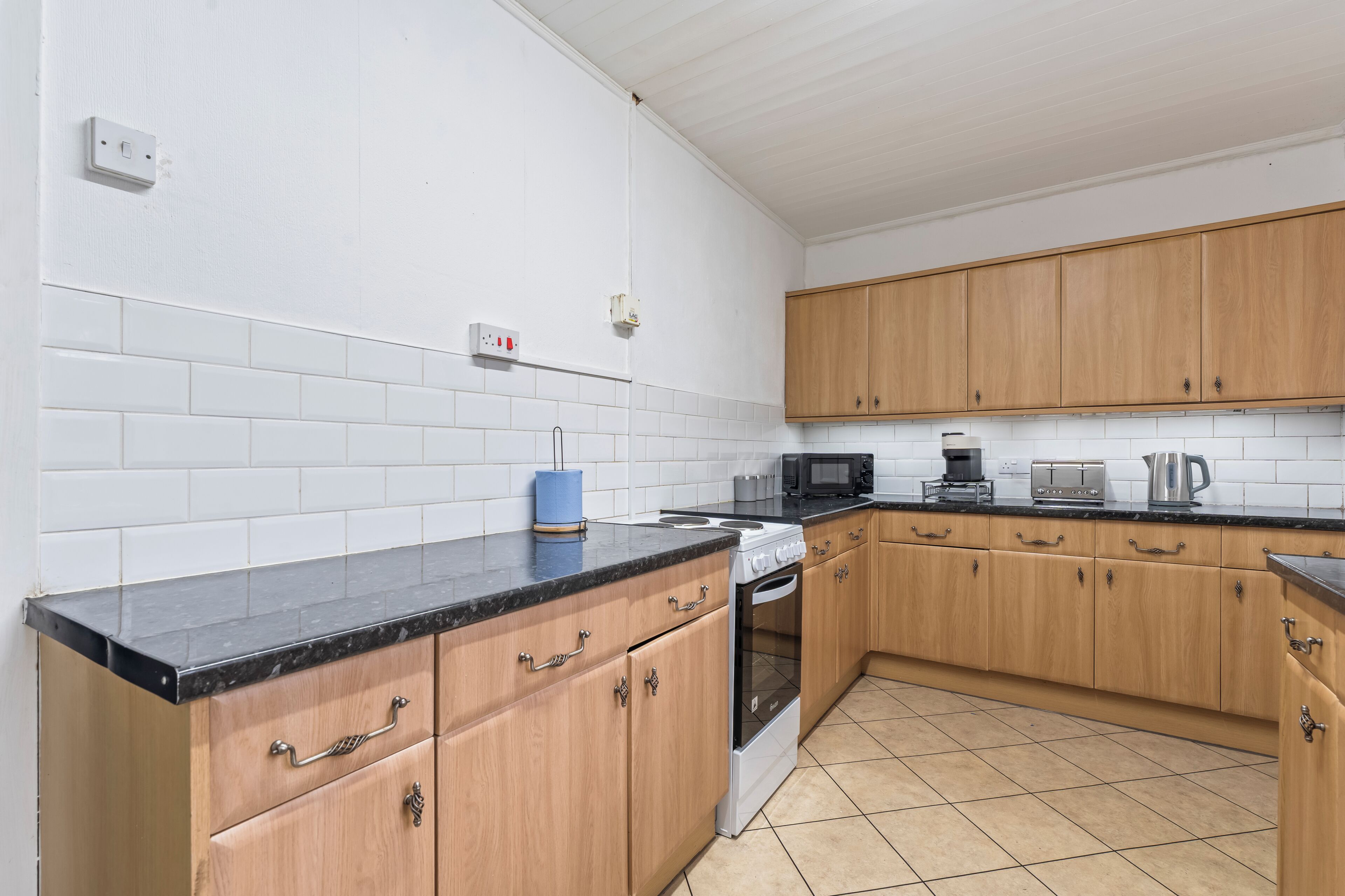 A kitchen with white tiled walls, wooden cabinets, a black countertop, and a stainless steel kettle.