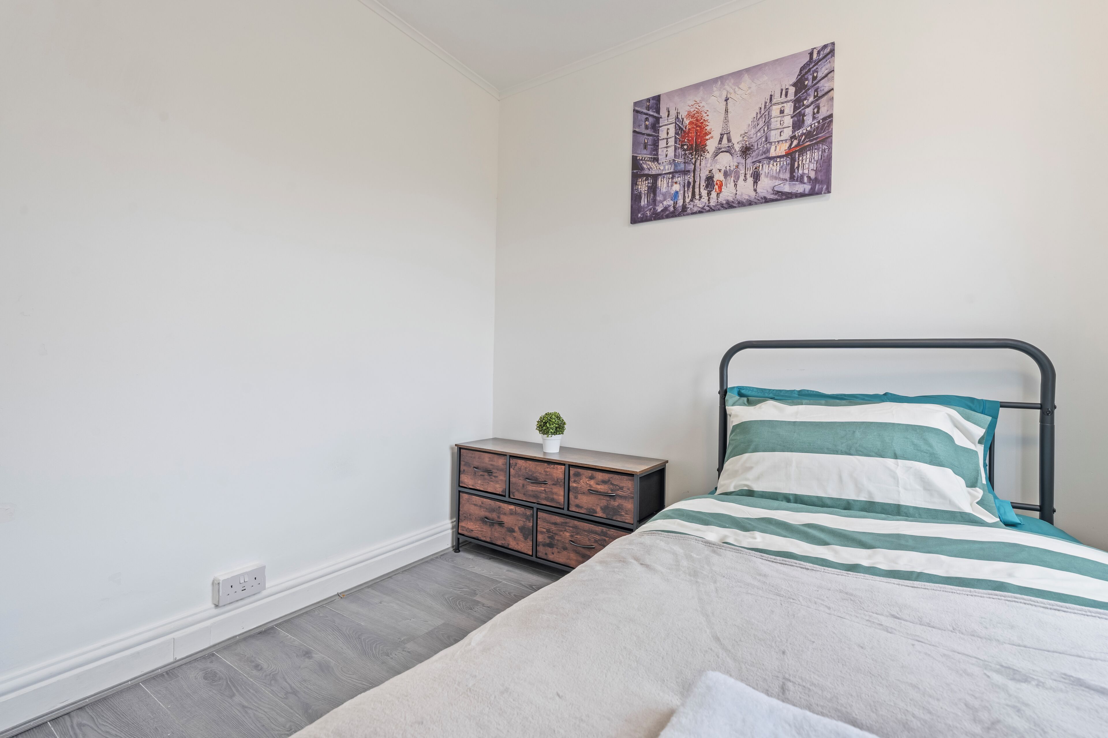 A bedroom with a bed, a dresser, a painting, and a potted plant.