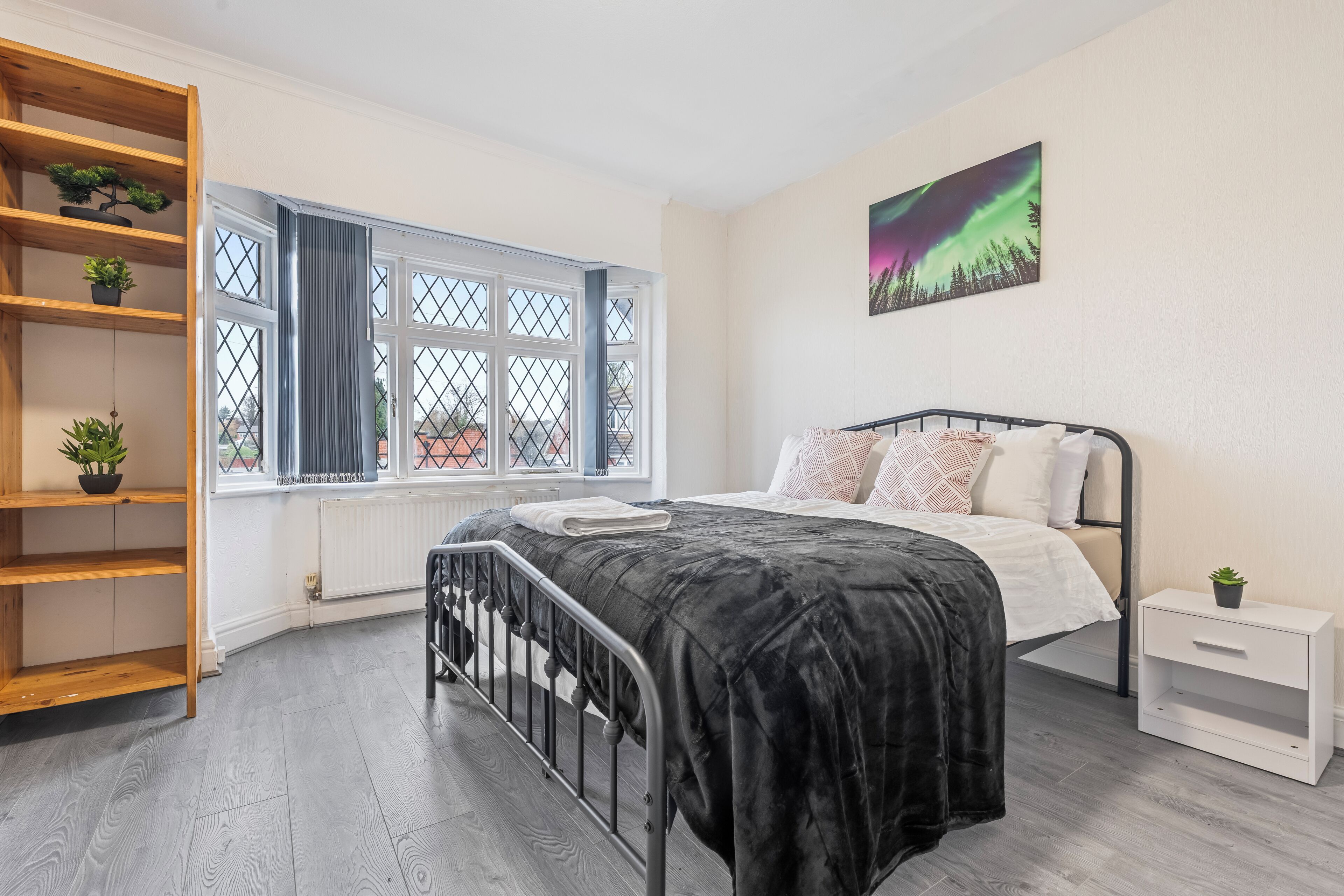 A bedroom with a bed, a window with a view, a shelf with plants, and a painting on the wall.