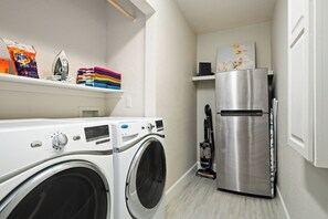 A compact laundry room with a front-loading washing machine, a stainless steel refrigerator, and a shelf with laundry detergent and folded towels. - Spacious 5-bedroom house fully Furnished Private Pool – Extended Stays Welcome (Scottsdale)