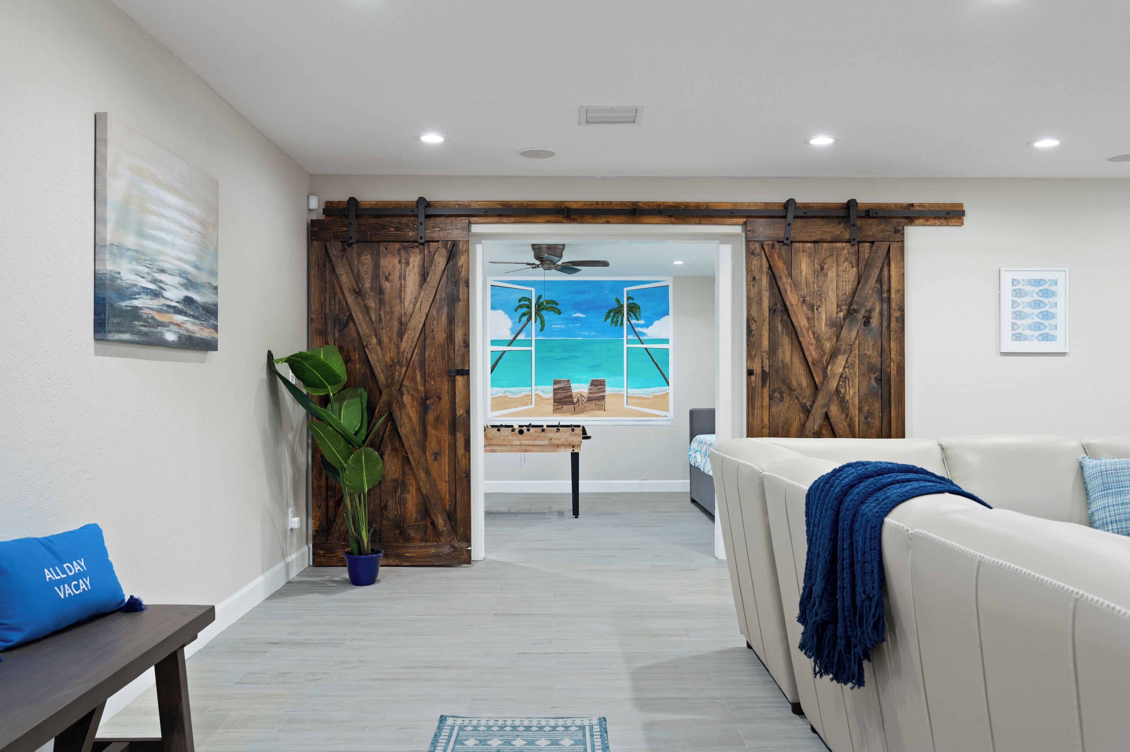 Un salón con vistas a una playa a través de puertas correderas, un sofá blanco, un cojín azul y una planta grande.