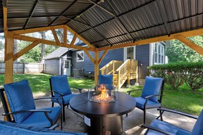 An outdoor patio with a round table and four chairs, a fire pit in the center, and a covered area with a metal roof.