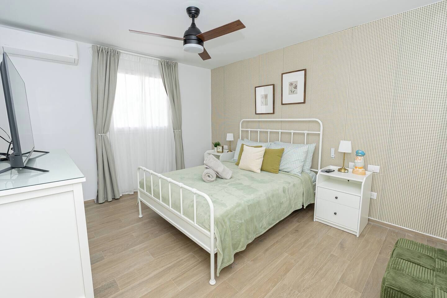 Un dormitorio con cama, mesitas de noche, ventilador de techo y ventana con cortinas.