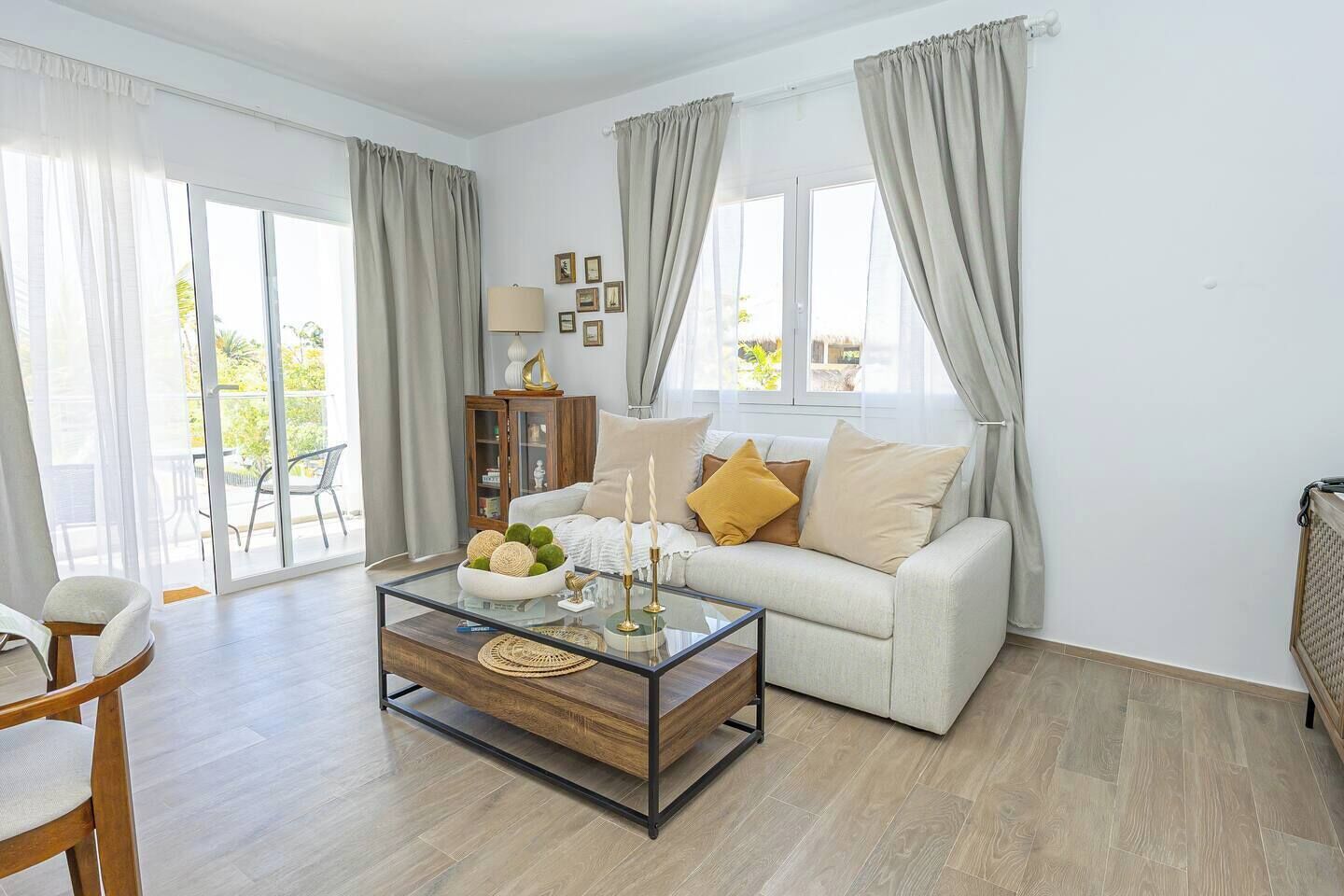 Una sala de estar moderna con una mesa de centro de vidrio, un sofá con cojines, un mueble de madera y vistas a través de ventanas amplias.