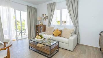 Ein modernes Wohnzimmer mit einem Glastisch, einer Couch mit Kissen, einer Holzkommode und Blick durch große Fenster.
