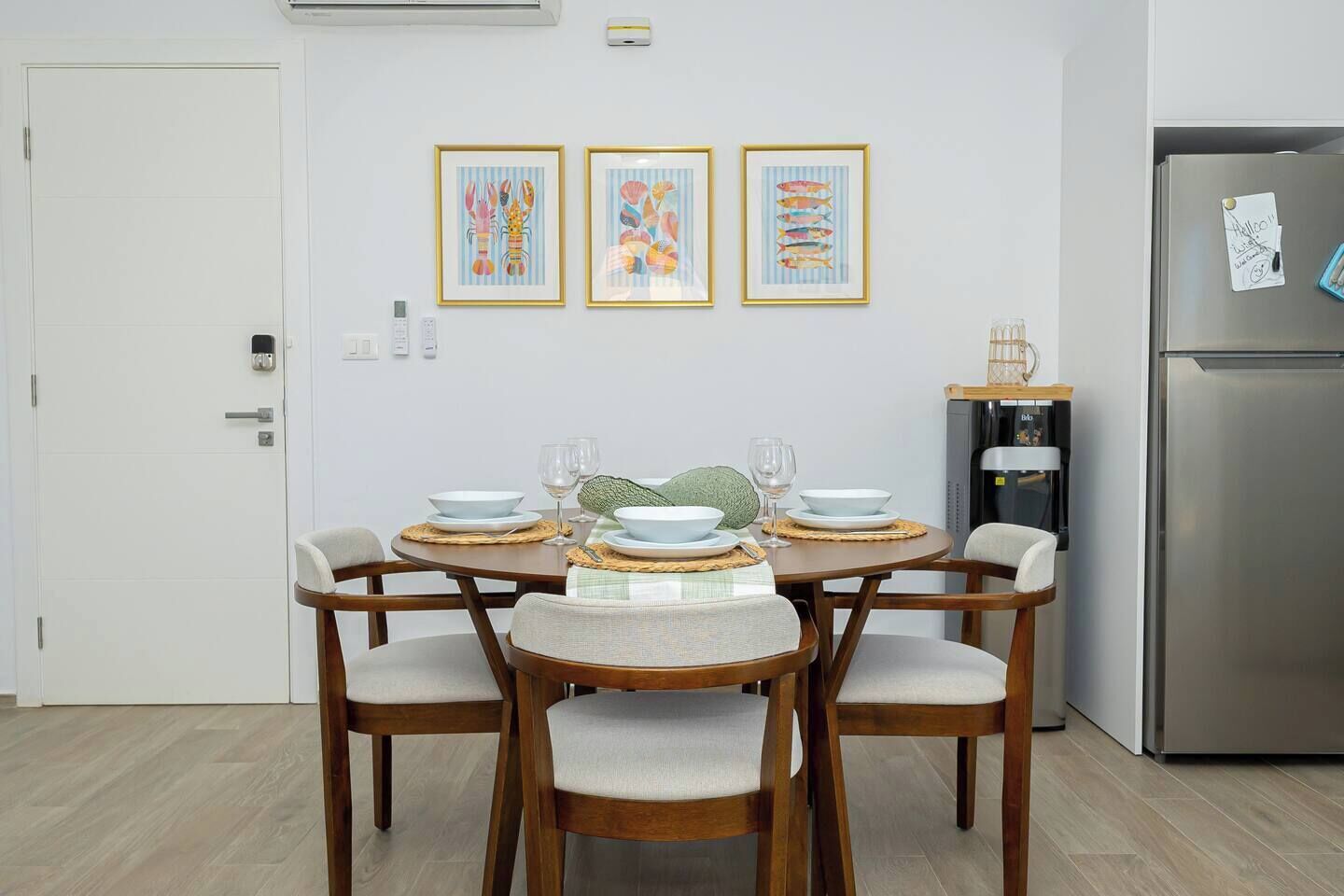 Ein Essbereich mit einem Tisch, gedeckt für vier Personen, umgeben von Holzstühlen mit gepolsterten Sitzen. An der Wand über dem Tisch hängen drei gerahmte Kunstwerke.