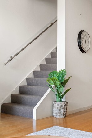 Eine Treppe mit grauen Teppichstufen, einem Metallgeländer, einer Uhr an der Wand und einem Topf mit breitblättrigem Grün.