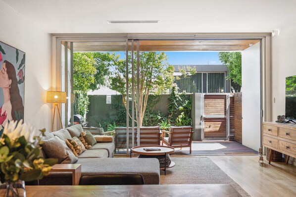 A modern living room with a sofa, coffee table, and chairs, a television, and a sliding glass door leading to a balcony with plants and a view of the outside.
