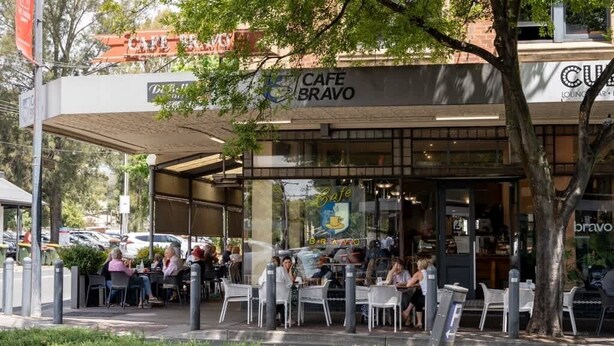 An outdoor cafe with tables and chairs, a covered seating area, and a building with a cafe sign. - Arcady, Norwood Townhouse Near The Parade and City (Norwood)
