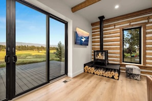 A modern interior with a fireplace, a large window, and a view of a green landscape. - The Willow House - Hot Tub w/ Views near Glacier (Columbia Falls)