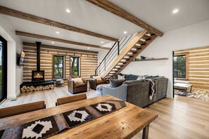 A log cabin interior with a fireplace, wooden beams, a staircase, and a sofa. - The Willow House - Hot Tub w/ Views near Glacier (Columbia Falls)
