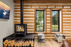A log cabin interior with a wood-burning stove, a window with a view of the outdoors, and a stack of firewood. - The Willow House - Hot Tub w/ Views near Glacier (Columbia Falls)