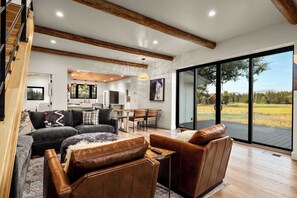 A spacious living area with a large sliding glass door leading to an outdoor view, a kitchen in the background, and a staircase on the left. - The Willow House - Hot Tub w/ Views near Glacier (Columbia Falls)