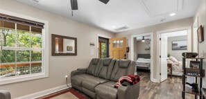 A spacious living room with a gray leather sofa, a large window, and a door leading to a bedroom.