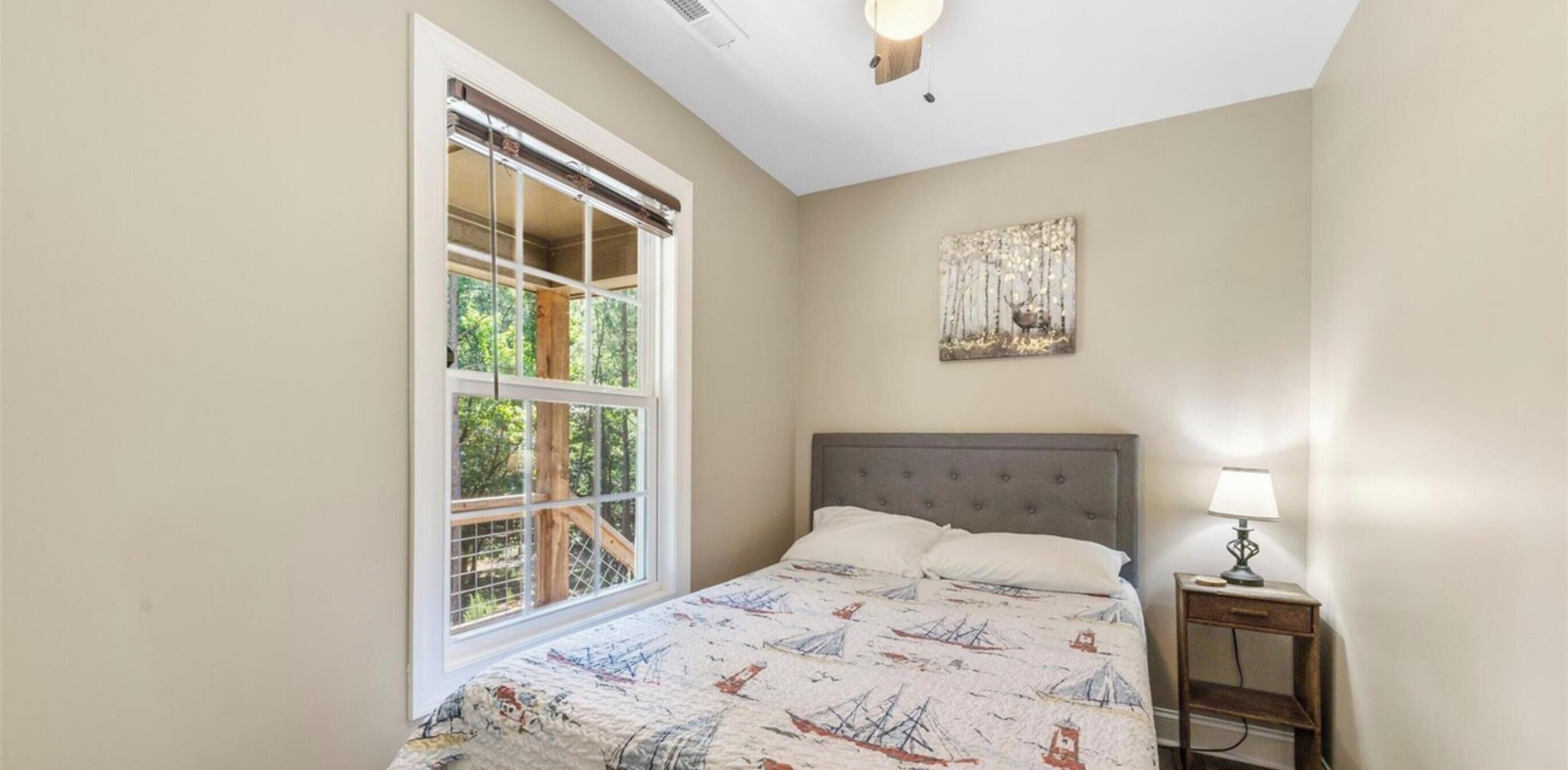 A bedroom with a bed, a nightstand, a lamp, a window with blinds, and a wall-mounted artwork.