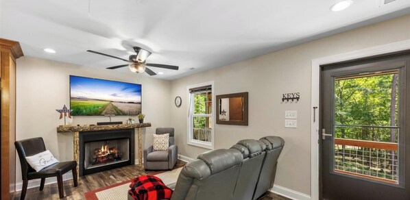 A living room with a fireplace, a television, a ceiling fan, and a door with a view of greenery.