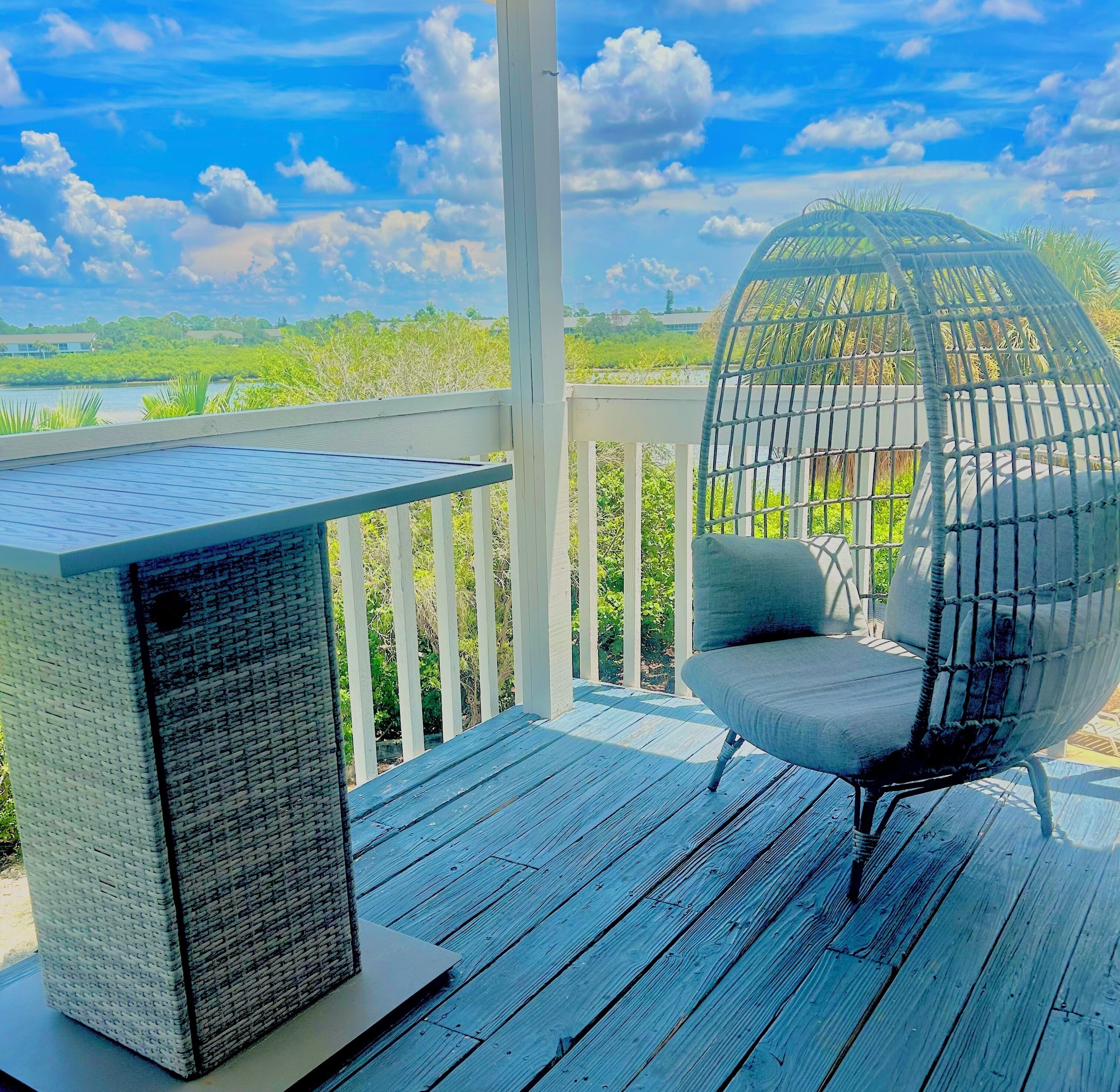 A deck with a woven chair and a table, overlooking a body of water and greenery.