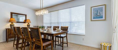 Un comedor con mesa de madera, sillas, una araña, ventana con persianas, un espejo y un cuadro en la pared.